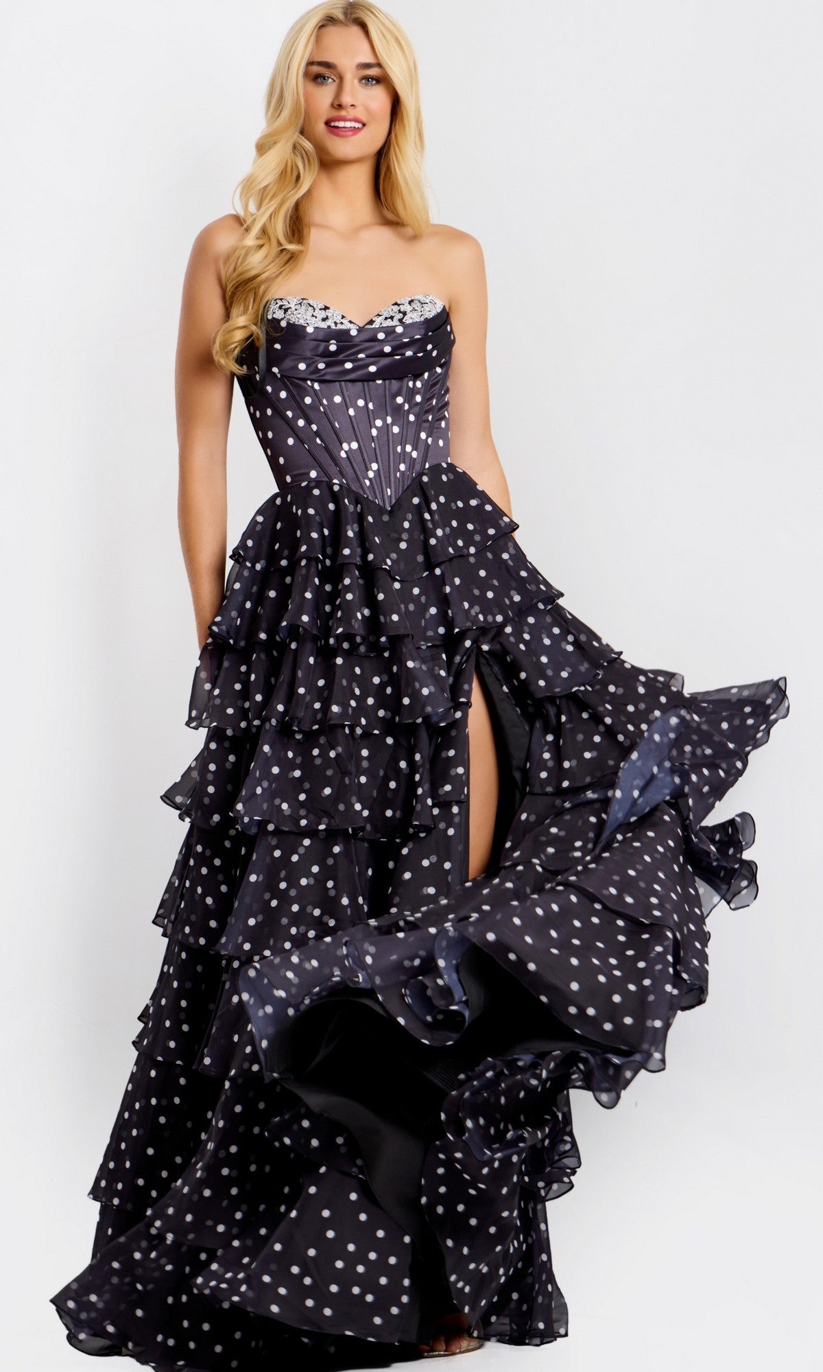 A woman poses against a plain background in the black Jovani 49087 long prom dress, featuring a strapless polka-dot design, ruffled layers, fitted bodice, and thigh-high slit—perfect for making a statement.